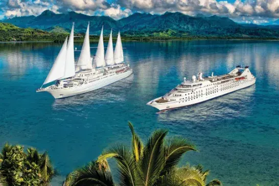 Two large cruise ships sailing on calm blue water—one with white sails and the other a modern liner without sails. Lush green palm trees and tropical vegetation are in the foreground, with green mountains and a partly cloudy sky in the background.