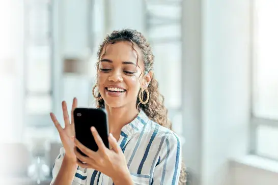 Person in striped shirt smiling at smartphone in a sunlit indoor space.