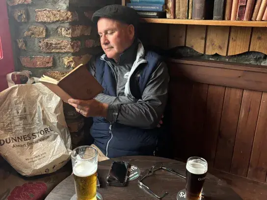 John Duggan reading a book at a small table in a cozy pub, with two pints of beer, glasses, and a bookshelf of old books in the background.