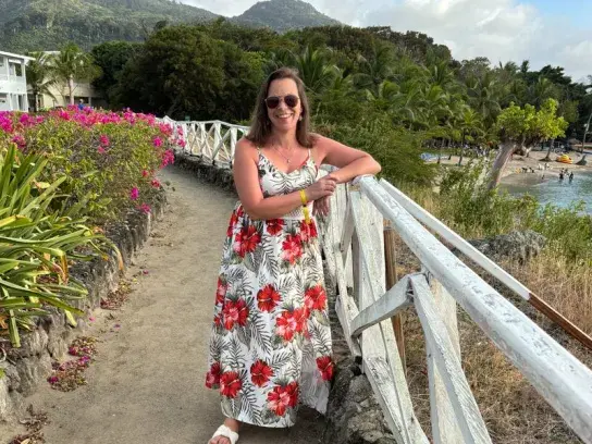 Dana standing beside a white railing along a scenic coastal path lined with tropical plants, with mountains, palm trees, and a beach visible in the background.