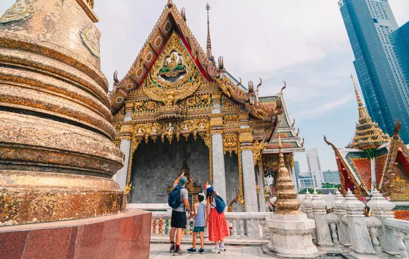 Three tourists with backpacks stand in front of an ornate golden temple, with modern skyscrapers in the background.