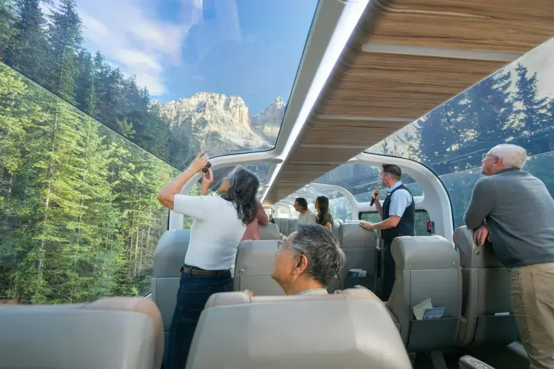 Scenic train interior with glass dome windows showing stunning mountain peaks and dense forest, passengers taking photos and enjoying panoramic views.