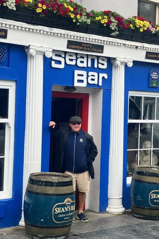 John standing in the doorway of Sean's Bar, a bright blue pub with white columns, flower boxes, and whiskey barrels at the entrance.
