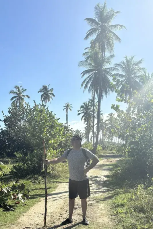 John holding a walking stick on a sandy path surrounded by lush tropical vegetation and tall palm trees under a bright blue sky.