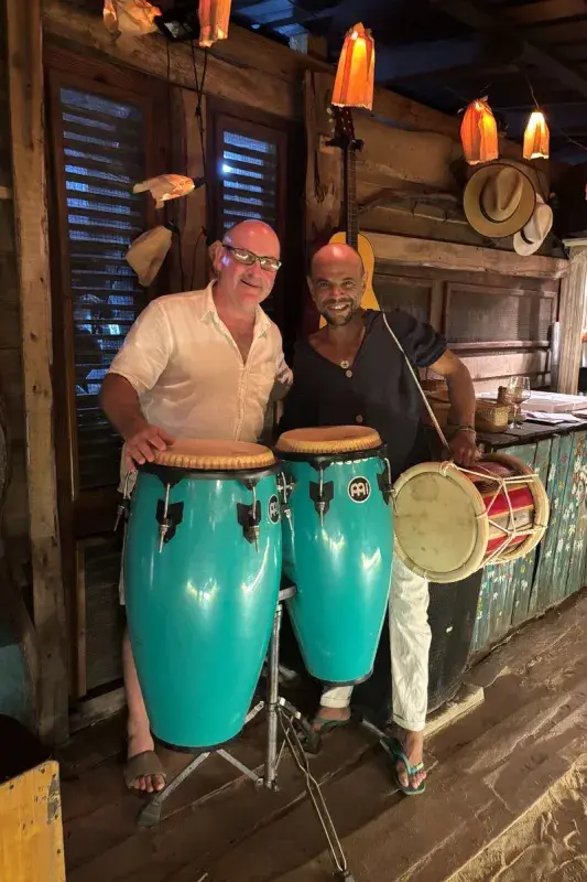Two people standing beside turquoise conga drums and a traditional drum inside a rustic, wood-paneled venue decorated with hanging lights, hats, and a guitar.