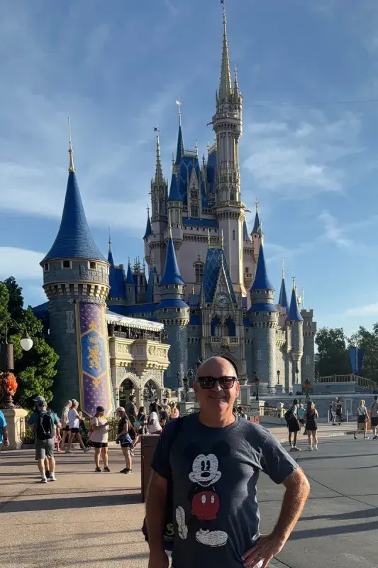 John standing in front of Cinderella Castle at a Disney theme park, with blue spires, surrounding walkways, and park visitors visible in the foreground.