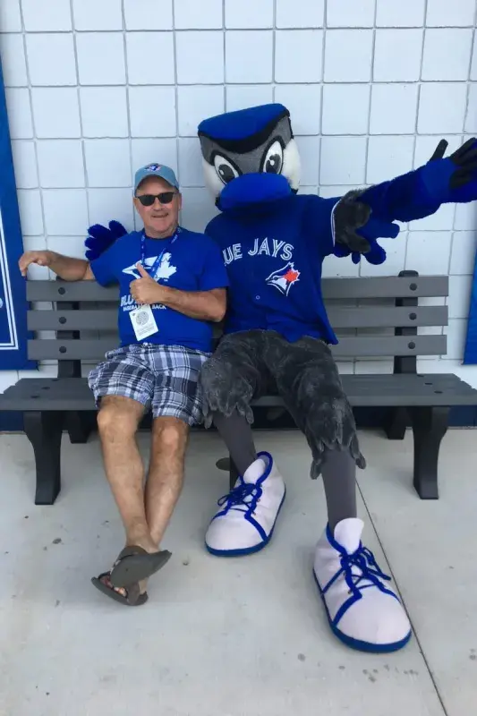 John seated on a bench beside the Toronto Blue Jays mascot, posing for a photo outside a stadium concourse.