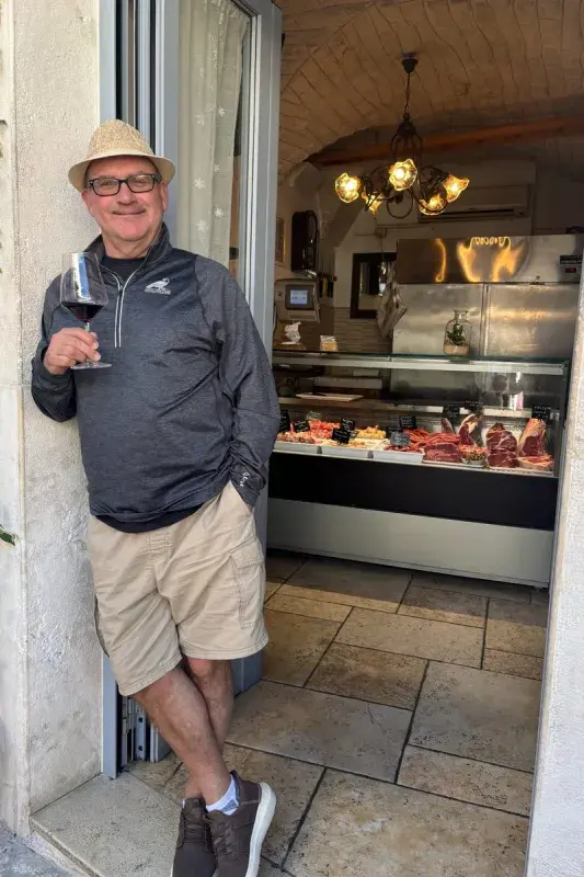 John holding a glass of red wine while standing in the doorway of a specialty food shop, with a display case of meats and warm interior lighting visible behind them.